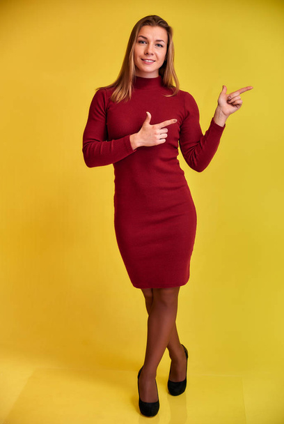 Full length vertical portrait of a pretty fitness blonde girl with a beautiful smile in a burgundy dress on a yellow background. Cute looks at the camera, standing straight. - Foto, afbeelding