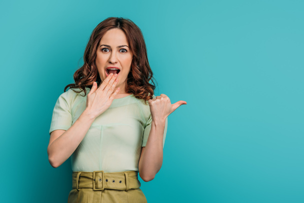 shocked woman pointing with thumb and looking at camera on blue background - Фото, изображение