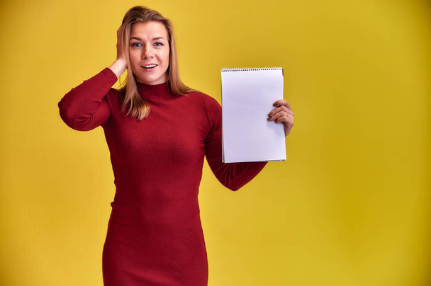 Portrait of a pretty blonde girl with a beautiful smile and excellent teeth in a burgundy dress with a folder in her hands on a yellow background. Cute looks at the camera, standing straight. - Foto, Imagen