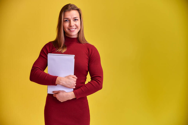 Portrait of a pretty blonde girl with a beautiful smile and excellent teeth in a burgundy dress with a folder in her hands on a yellow background. Cute looks at the camera, standing straight. - Zdjęcie, obraz