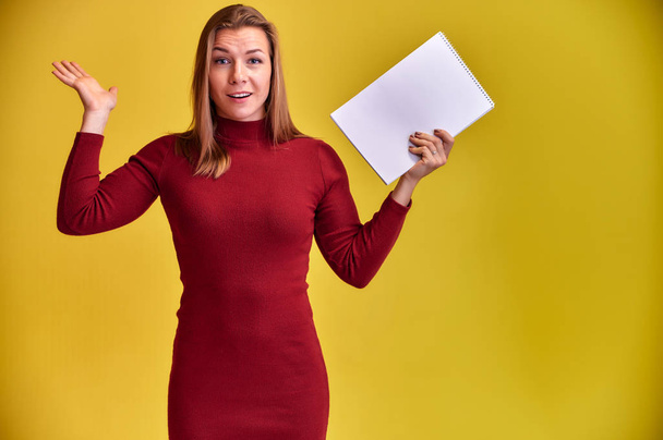 Portrait of a pretty blonde girl with a beautiful smile and excellent teeth in a burgundy dress with a folder in her hands on a yellow background. Cute looks at the camera, standing straight. - 写真・画像