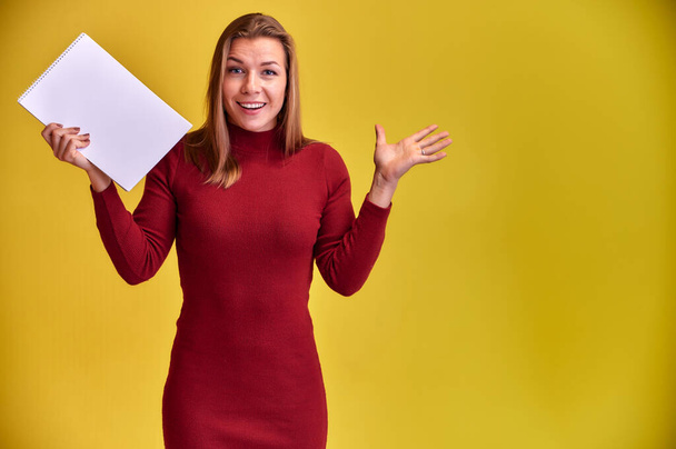 Portrait of a pretty blonde girl with a beautiful smile and excellent teeth in a burgundy dress with a folder in her hands on a yellow background. Cute looks at the camera, standing straight. - Fotografie, Obrázek