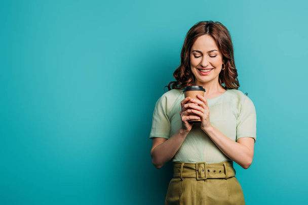 happy girl smiling while holding coffee to go on blue background - Valokuva, kuva