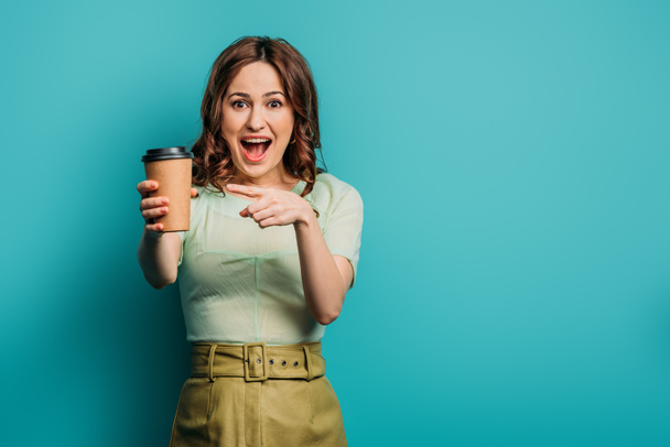 cheerful girl pointing with finger at paper cup on blue background - Φωτογραφία, εικόνα
