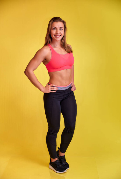Vertical portrait of a pretty fitness blonde girl with a beautiful smile, excellent teeth and a bodybuilder figure in a tracksuit on a yellow background. Cute looks at the camera, standing straight. - 写真・画像
