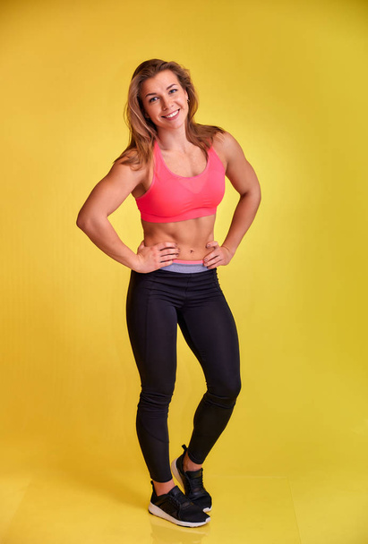 Vertical portrait of a pretty fitness blonde girl with a beautiful smile, excellent teeth and a bodybuilder figure in a tracksuit on a yellow background. Cute looks at the camera, standing straight. - Foto, imagen