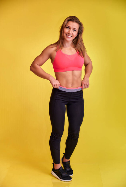 Vertical portrait of a pretty fitness blonde girl with a beautiful smile, excellent teeth and a bodybuilder figure in a tracksuit on a yellow background. Cute looks at the camera, standing straight. - Fotó, kép