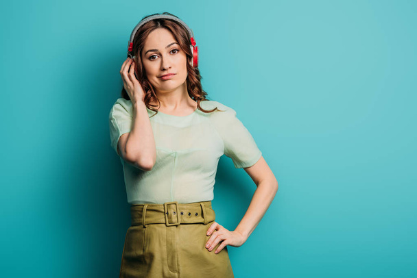 skeptical girl in wireless headphones standing with hand on hip on blue background - Photo, Image