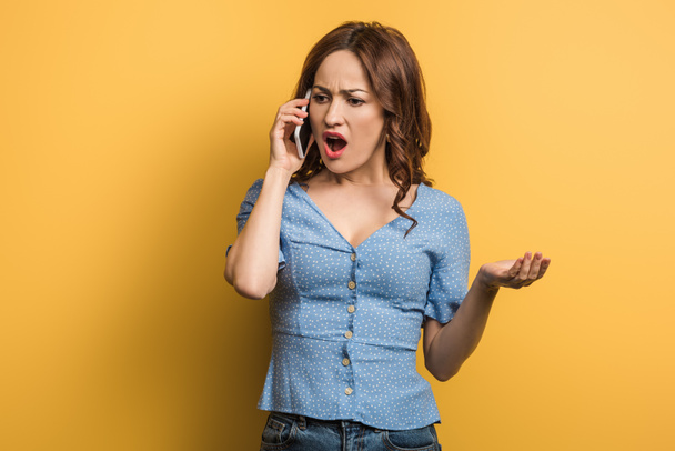 irritated woman standing with open arm while talking on smartphone on yellow background - Фото, зображення