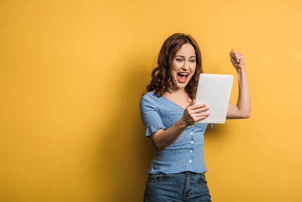 excited woman showing winner gesture while holding digital tablet on yellow background - Foto, Bild