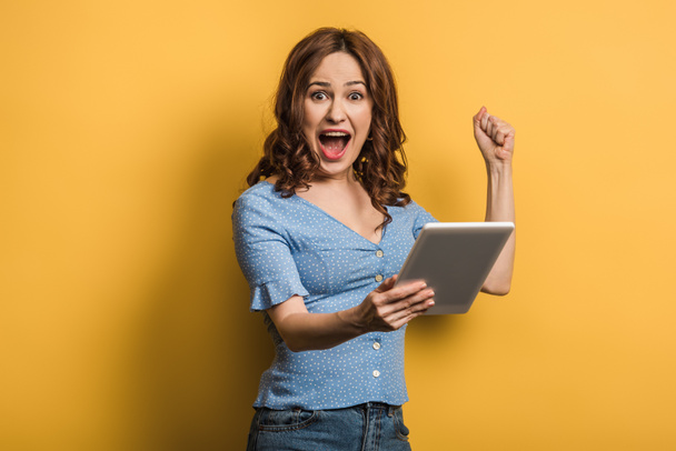 excited woman showing winner gesture while holding digital tablet on yellow background - Фото, зображення