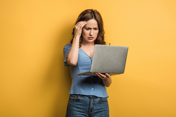 thoughtful girl holding hand near head while looking at laptop on yellow background - Φωτογραφία, εικόνα