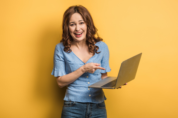 cheerful girl looking at camera while pointing with finger at laptop on yellow background - Фото, изображение