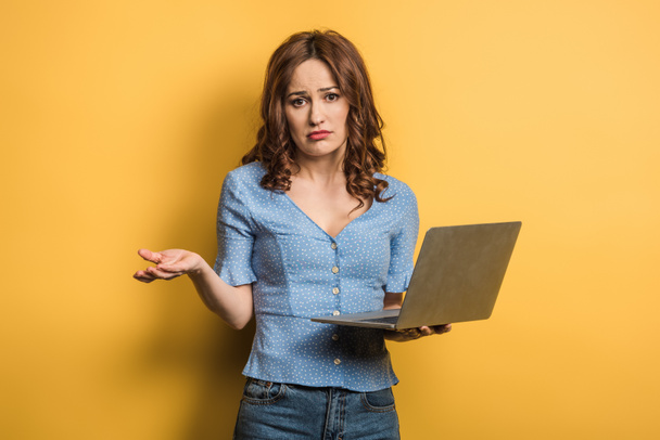 chica confundida mostrando gesto encogiéndose de hombros mientras sostiene el ordenador portátil sobre fondo amarillo
 - Foto, Imagen