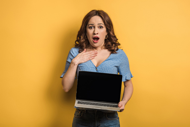 shocked woman touching chest while holding laptop with blank screen on yellow background - 写真・画像