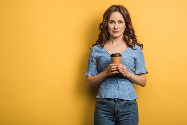 smiling young woman holding coffee to go while looking at camera on yellow background - Photo, image