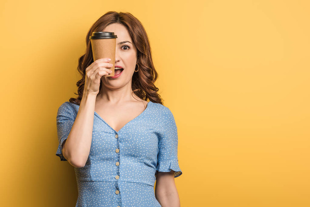 cheerful woman covering eye with paper cup on yellow background - Foto, Imagem