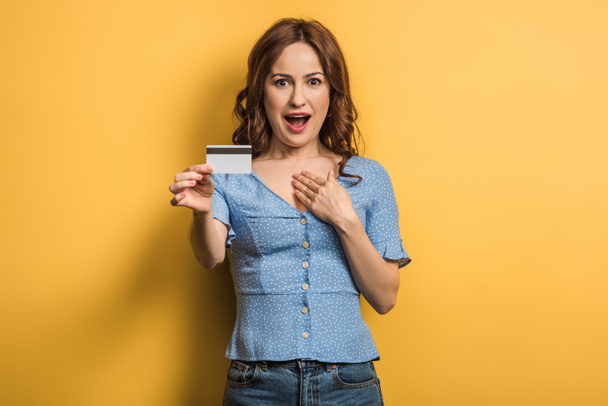 surprised woman holding credit card while looking at camera on yellow background - Photo, Image