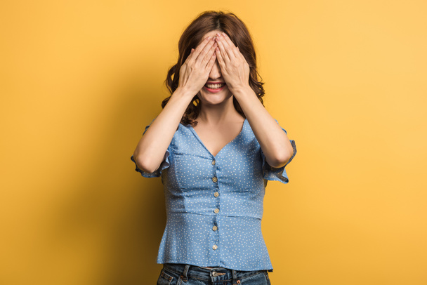 cheerful girl covering eyes with hands on yellow background - Fotó, kép