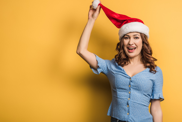 happy girl touching santa hat while looking at camera on yellow background - Foto, Imagem