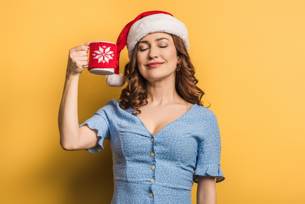 pleased girl in santa hat holding cup of warm drink with closed eyes on yellow background - Zdjęcie, obraz