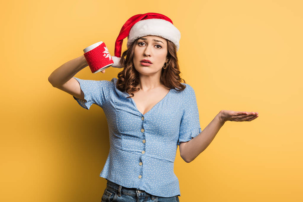 upset girl in santa hat holding empty cup on yellow background - Photo, Image
