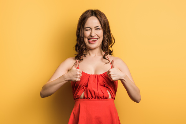 cheerful girl showing thumbs up, winking and sticking out tongue on yellow background - Foto, imagen