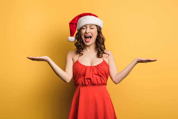 gelukkig meisje in santa hoed lachen met open armen op gele achtergrond - Foto, afbeelding