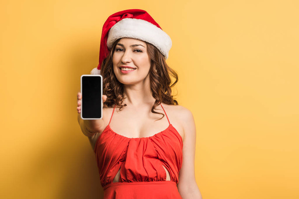 χαμογελαστό κορίτσι στο καπέλο santa δείχνει smartphone με λευκή οθόνη στο κίτρινο φόντο - Φωτογραφία, εικόνα