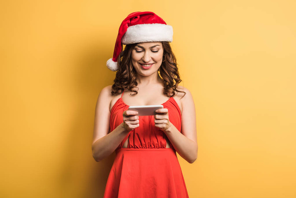 cheerful girl in santa hat messaging on smartphone on yellow background - Photo, Image