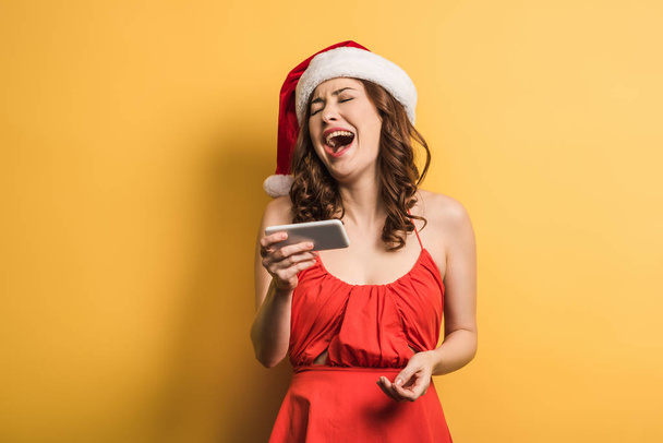 cheerful girl in santa hat laughing during video call on yellow background - Фото, зображення