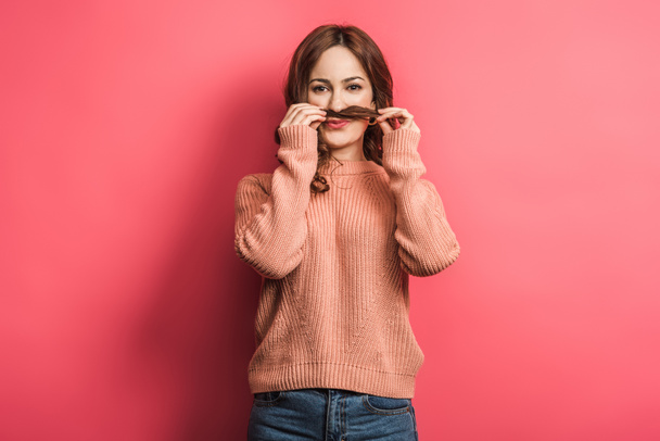 tricky girl holding hair above lip while imitating mustache on pink background - Photo, Image