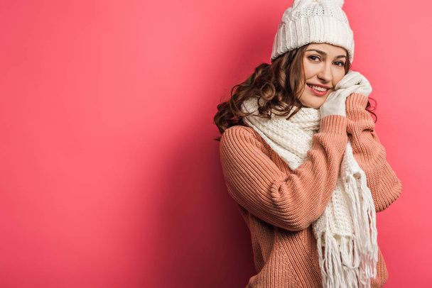 cold, smiling girl in warm hat and scarf standing with clenched hands on pink background - Valokuva, kuva