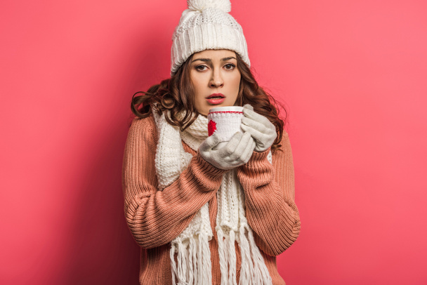 cold girl in warm hat and scarf holding cup with warming drink on pink background - Foto, imagen
