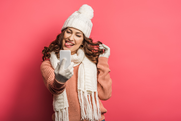 tricky girl in warm hat and scarf sticking out tongue during video call on smartphone on pink background - Photo, Image