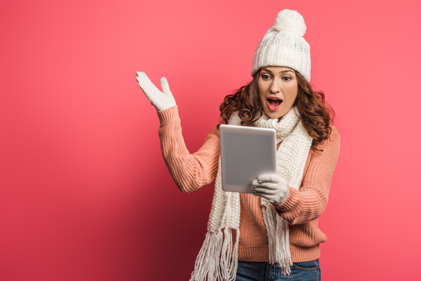 surprised girl in warm hat and scarf looking at digital tablet on pink background - Valokuva, kuva