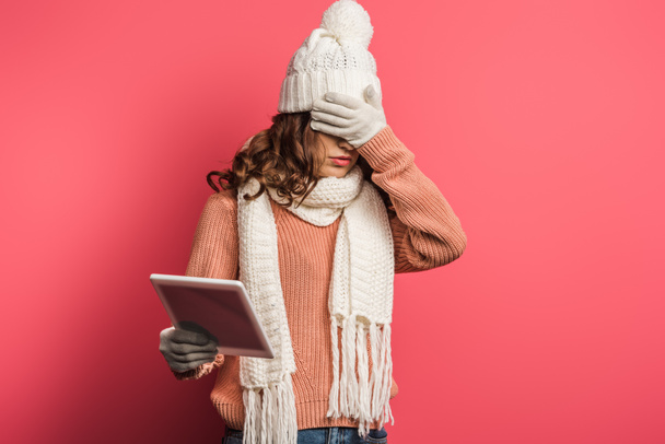 upset girl in warm hat and scarf holding digital tablet and covering face with hand on pink background - Фото, зображення