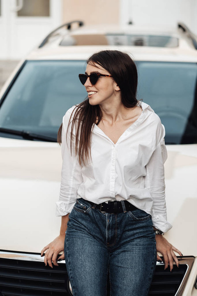 Close Up Retrato de uma elegante jovem mulher vestida de jeans e camisa branca posando perto do carro, senhora de negócios, conceito de poder da mulher
 - Foto, Imagem