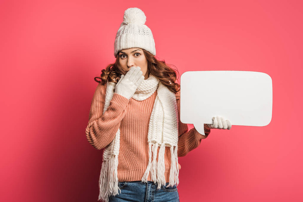 shocked girl in warm hat and scarf covering mouth while holding speech bubble on pink background - Фото, изображение