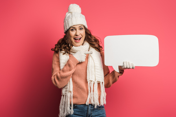 smiling girl in warm hat and scarf pointing with finger at speech bubble on pink background - Fotoğraf, Görsel