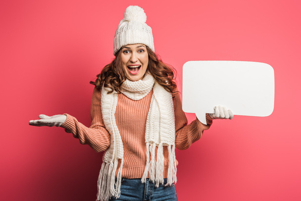 cheerful girl in warm hat and scarf holding speech bubble on pink background - Photo, Image