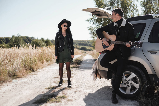 Szczęśliwa para podróżująca ubrana w czarne stylowe ubrania Ciesząca się wycieczką samochodową po polu, Człowiek gra na gitarze, Koncepcja wakacji - Zdjęcie, obraz