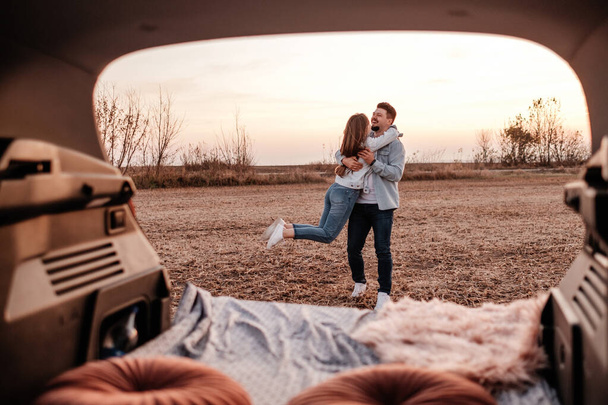 Joven pareja feliz vestida como en camisa blanca y pantalones vaqueros sentados en su nuevo baúl de coche, hermoso atardecer en el campo, vacaciones y concepto de viaje
 - Foto, imagen