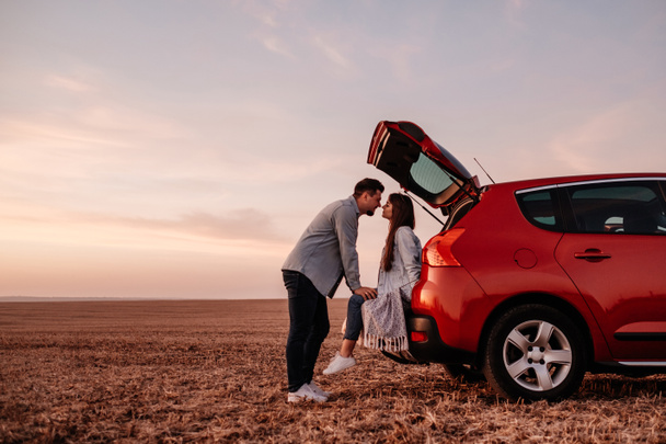 Młoda szczęśliwa para ubrana jak w białej koszuli i dżinsach siedzi na ich nowym bagażniku samochodu, piękny zachód słońca na polu, wakacje i podróży Concept - Zdjęcie, obraz