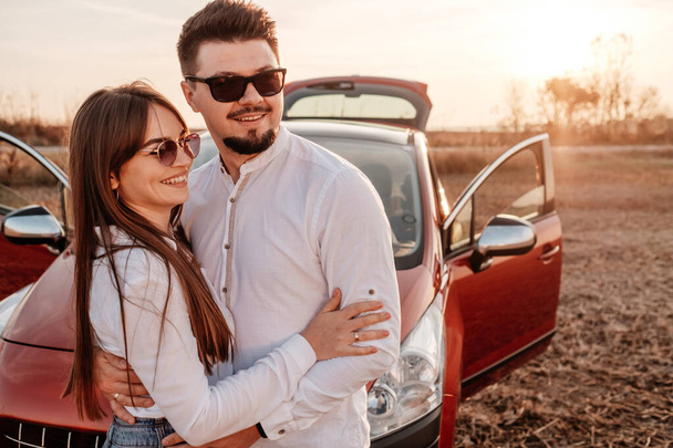 白いシャツを着た若い幸せなカップルと新しい車でロードトリップを楽しむジーンズ、フィールドでの美しい夕日、休暇と旅行のコンセプト - 写真・画像