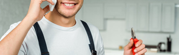 Panoramaaufnahme eines glücklichen Installateurs, der mit dem Smartphone spricht und eine Zange hält - Foto, Bild