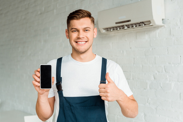 cheerful installer showing thumb up while holding smartphone with blank screen  - Foto, afbeelding