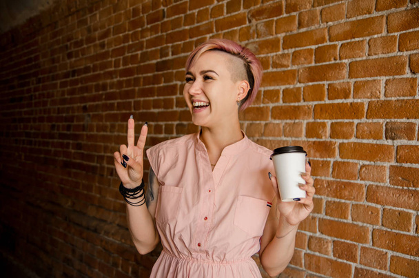 Молодая красивая улыбающаяся девушка держит бумажную чашку кофе на фоне кирпичной стены
 - Фото, изображение