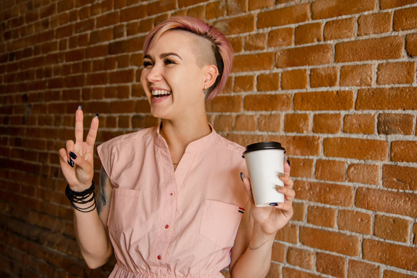 Young beautiful smiling girl holds paper cup of coffee on a brick wall background - Фото, изображение