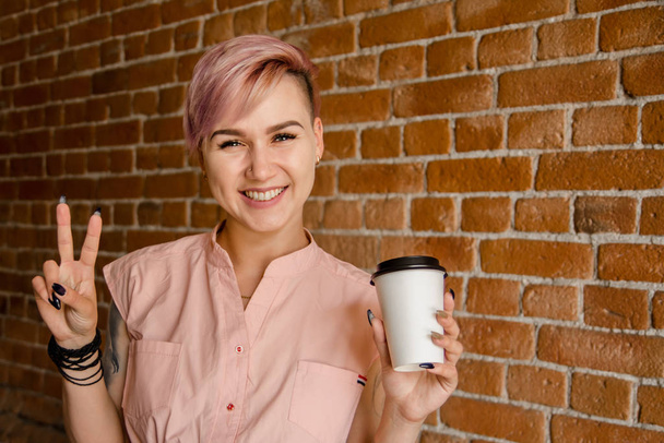 Young beautiful smiling girl holds paper cup of coffee on a brick wall background - Fotoğraf, Görsel